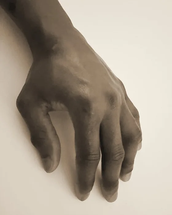 The artist’s left hand, resting on a pale table.